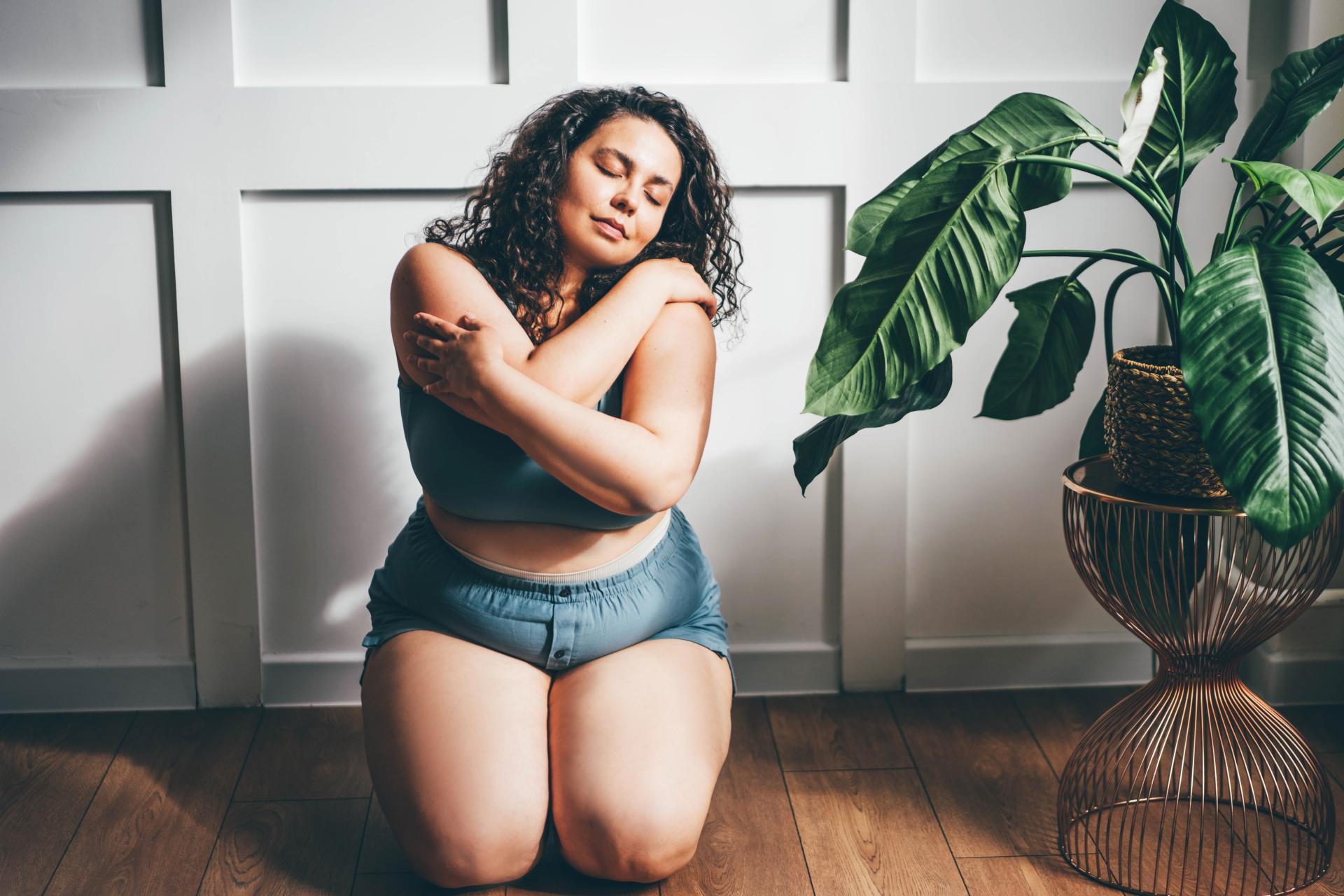 a photo of a woman kneeling on the floor near a lush green plant and lovingly hugging herself a photo of a woman kneeling on the floor near a lush green plant and lovingly hugging herself