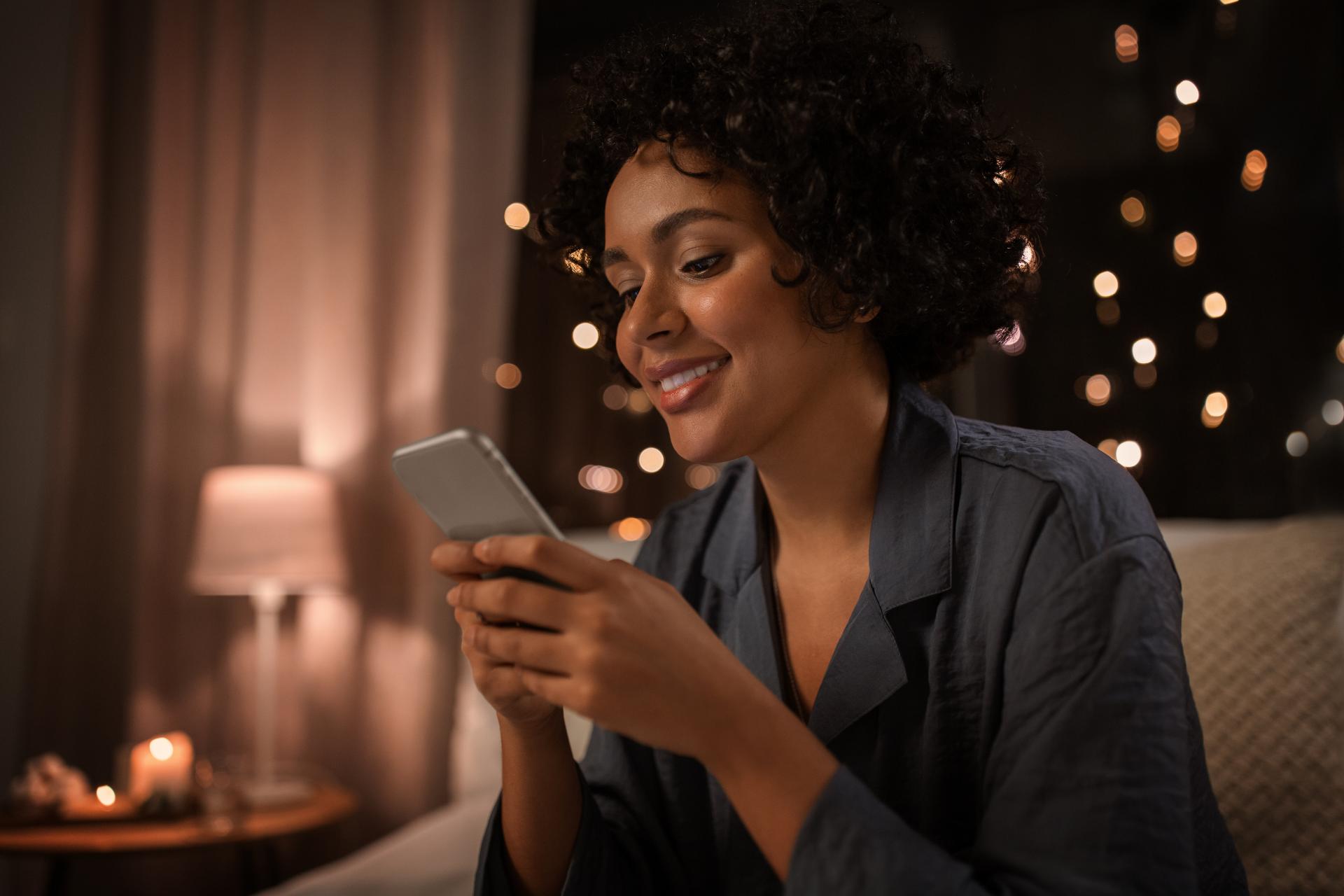 Woman smiling at her cell phone in bed at night with blurred background lighting Woman smiling at her cell phone in bed at night with blurred background lighting
