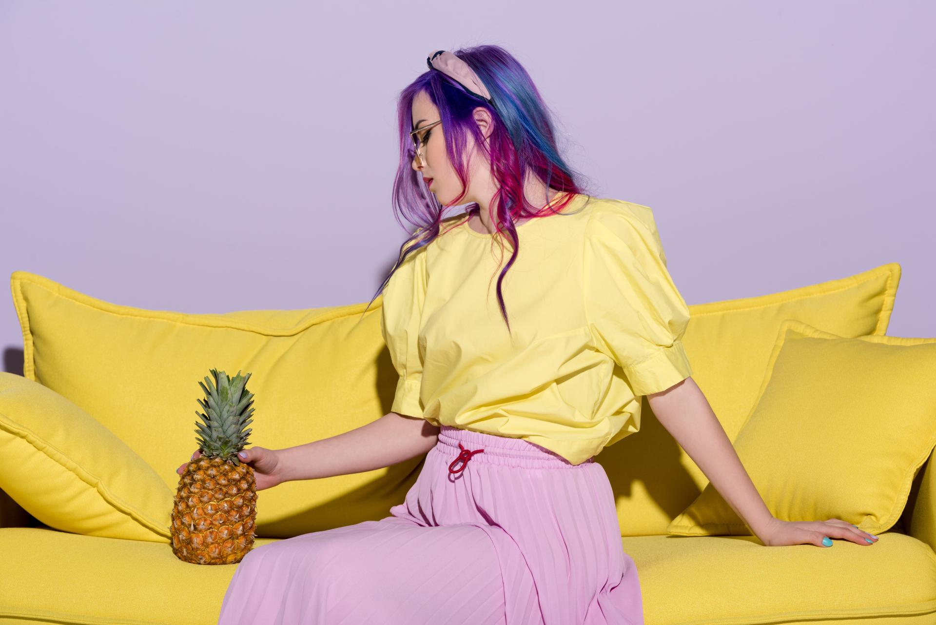 Woman with colorful hair, a yellow bouse, and pink skirt sitting with a pineapple on a yellow couch Woman with colorful hair, a yellow bouse, and pink skirt sitting with a pineapple on a yellow couch