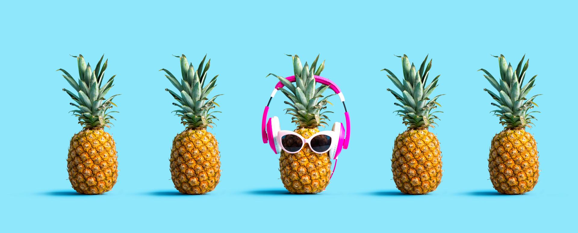 Row of fresh pineapples with one wearing sunglasses and headphones over a blue background Row of fresh pineapples with one wearing sunglasses and headphones over a blue background