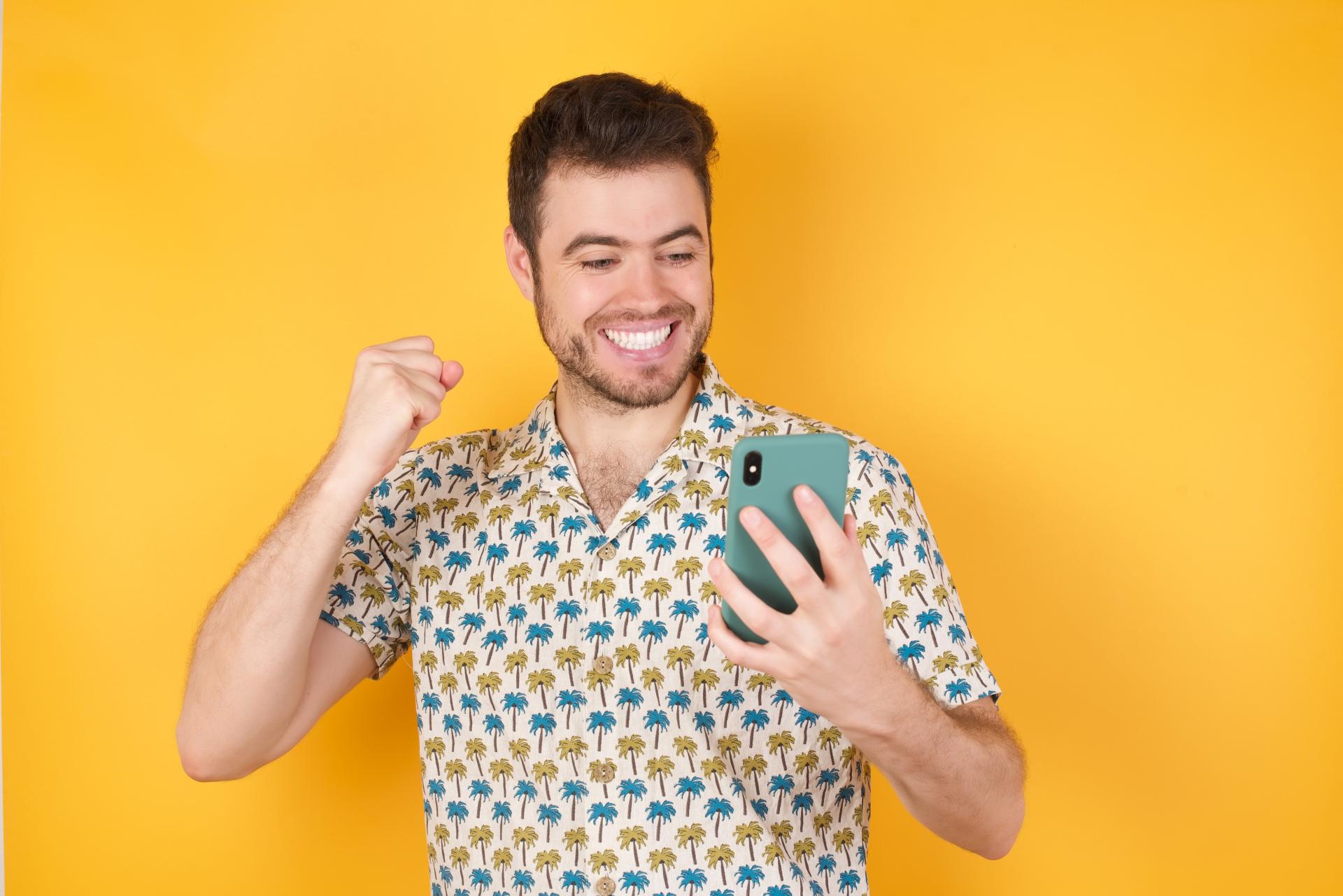 Excited man over a yellow background wearing a pineapple patterned shirt looking at his cell phone Excited man over a yellow background wearing a pineapple patterned shirt looking at his cell phone