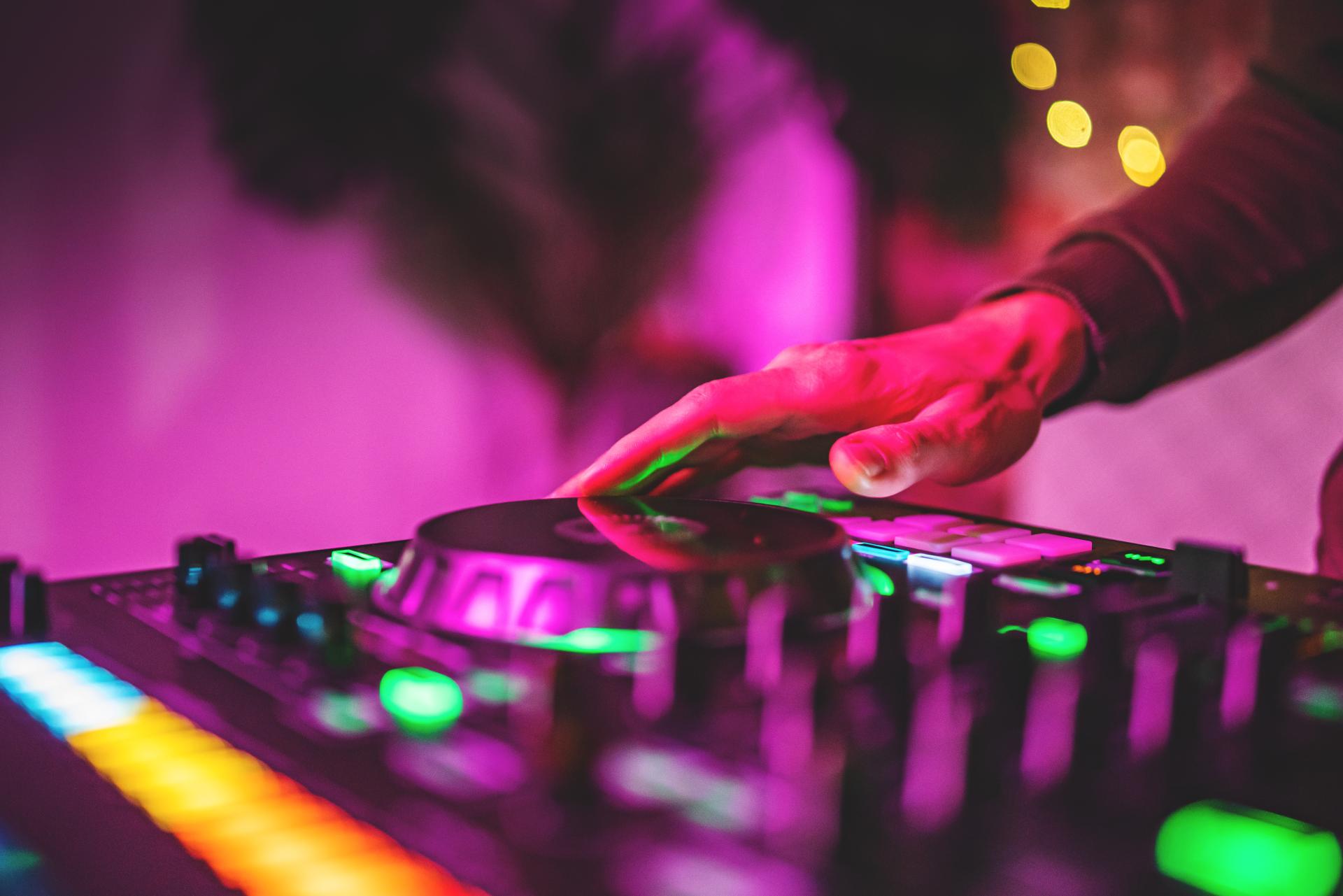 A hand working the controls of an illuminated DJ console mixer in a night club setting with purple l A hand working the controls of an illuminated DJ console mixer in a night club setting with purple l