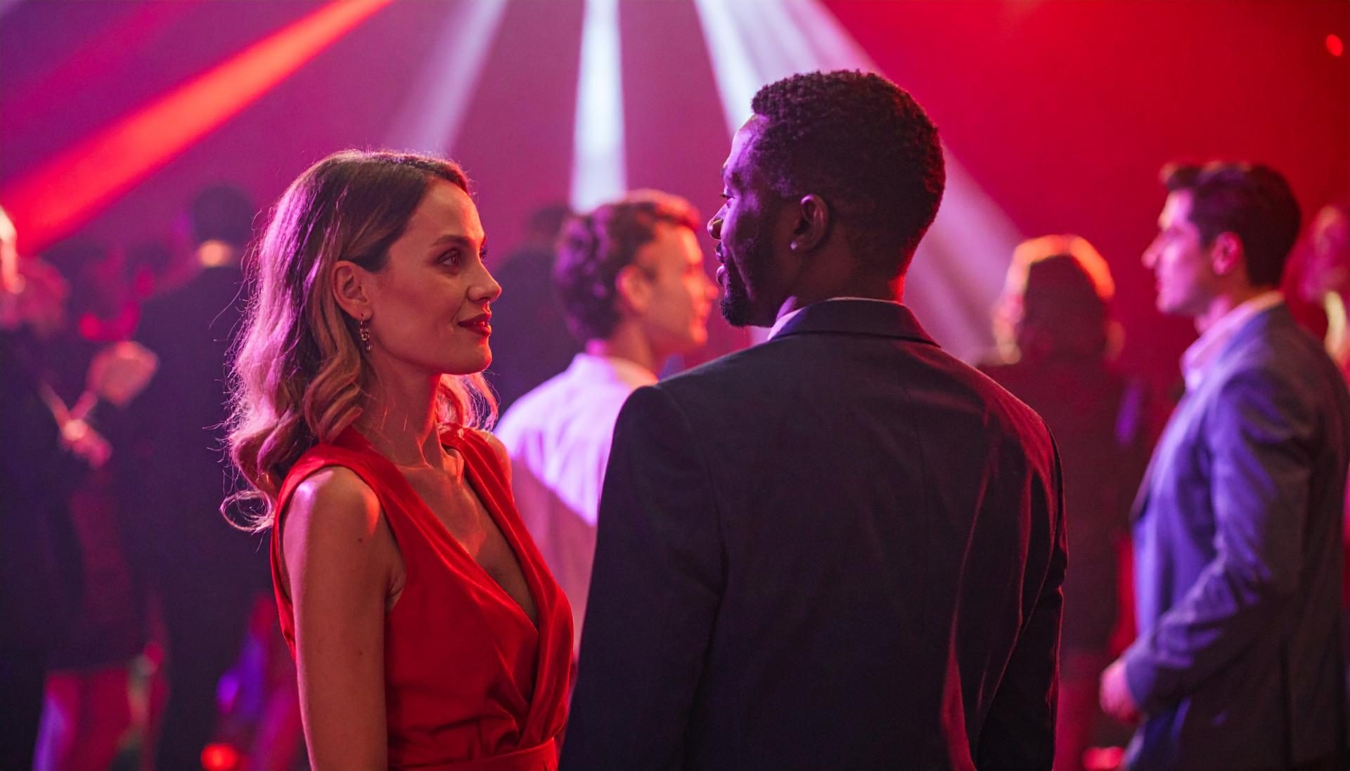 couple talking at nightclub Woman in a red dress smiles at a man in a dark suit on a crowded nightclub dance floor lit with red and purple lights.