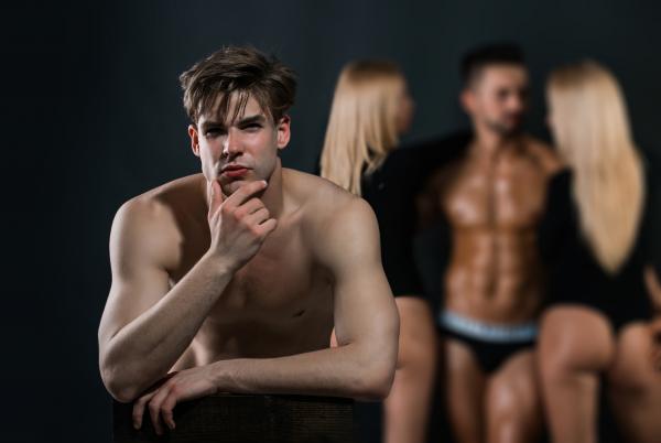 Male-presenting swinger in a thoughtful pose in front of a group of blurry people Male-presenting swinger in a thoughtful pose in front of a group of blurry people