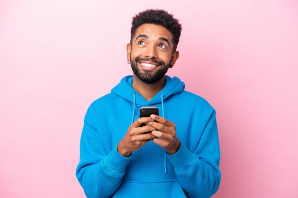 Hopeful smiling man with brown skin and a blue hoodie holding a smartphone over a pink background Hopeful smiling man with brown skin and a blue hoodie holding a smartphone over a pink background