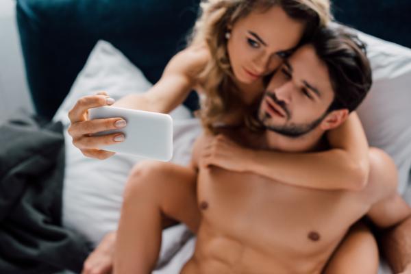 A photo of woman hugging boyfriend while taking selfie with smartphone on bed A photo of woman hugging boyfriend while taking selfie with smartphone on bed