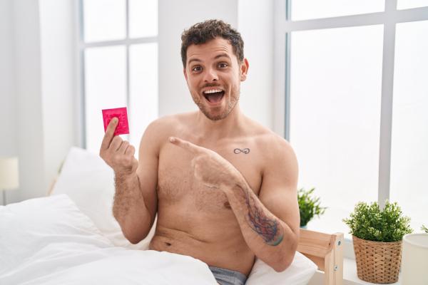 A photo of a Hispanic man holding condom in the bed smiling happy pointing with hand and finger A photo of a Hispanic man holding condom in the bed smiling happy pointing with hand and finger