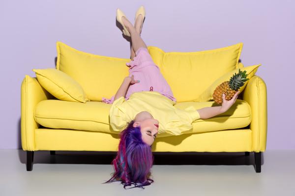 a photo of a woman with bisexual flag colored hair lying upside down on a yellow couch holding a pin a photo of a woman with bisexual flag colored hair lying upside down on a yellow couch holding a pin