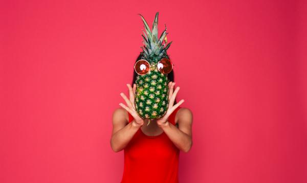femme person wearing red in front of a red background while covering their face with a pineapple femme person wearing red in front of a red background while covering their face with a pineapple