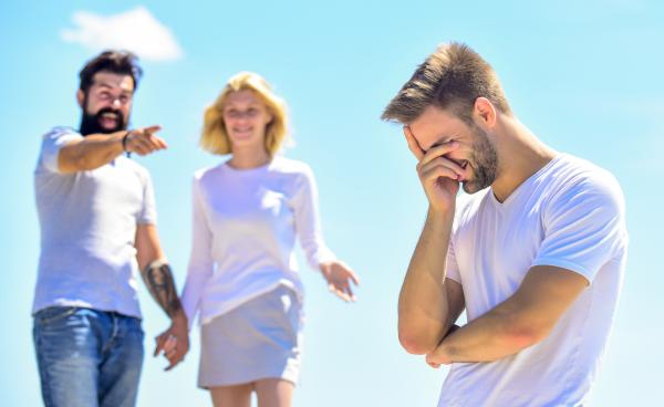 blonde man hiding face in his hand in front of a couple mocking and pointing at him blonde man hiding face in his hand in front of a couple mocking and pointing at him