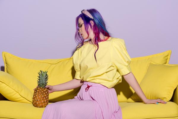 Woman with colorful hair, a yellow bouse, and pink skirt sitting with a pineapple on a yellow couch Woman with colorful hair, a yellow bouse, and pink skirt sitting with a pineapple on a yellow couch