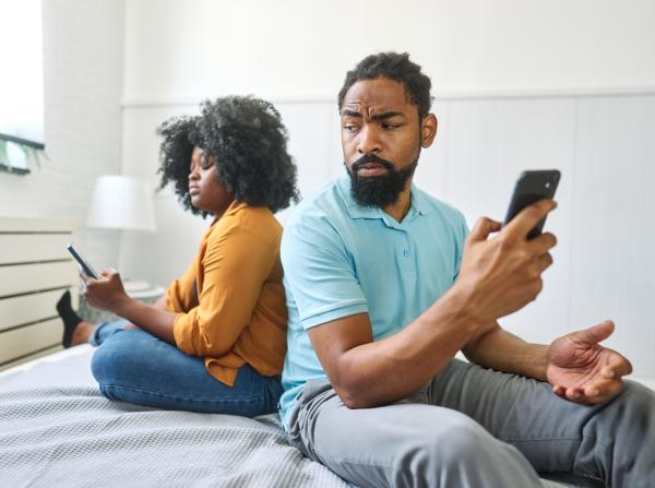 Couple sitting on a bed looking skeptical of each other while holding their mobile phones Couple sitting on a bed looking skeptical of each other while holding their mobile phones