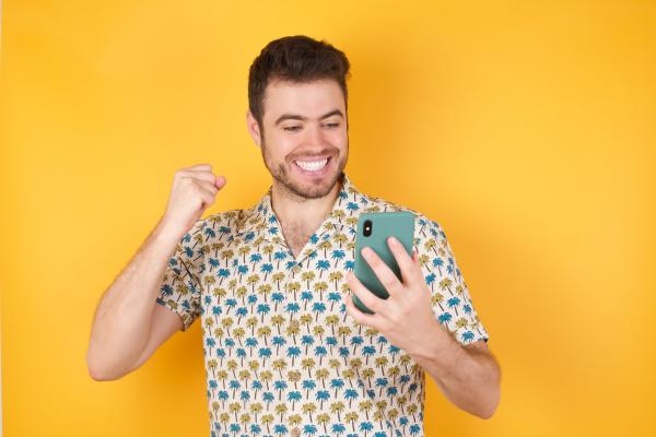 Excited man over a yellow background wearing a pineapple patterned shirt looking at his cell phone Excited man over a yellow background wearing a pineapple patterned shirt looking at his cell phone