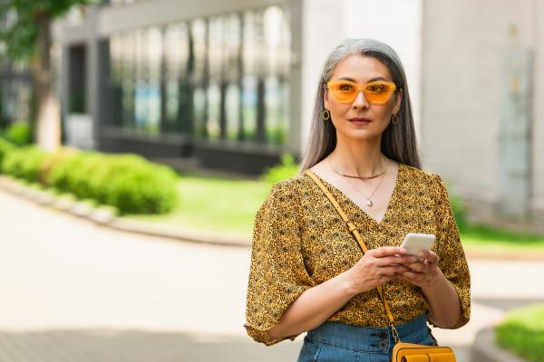 grey haired woman in patterned blouse and yellow sunglasses standing with smartphone outdoors grey haired woman in patterned blouse and yellow sunglasses standing with smartphone outdoors