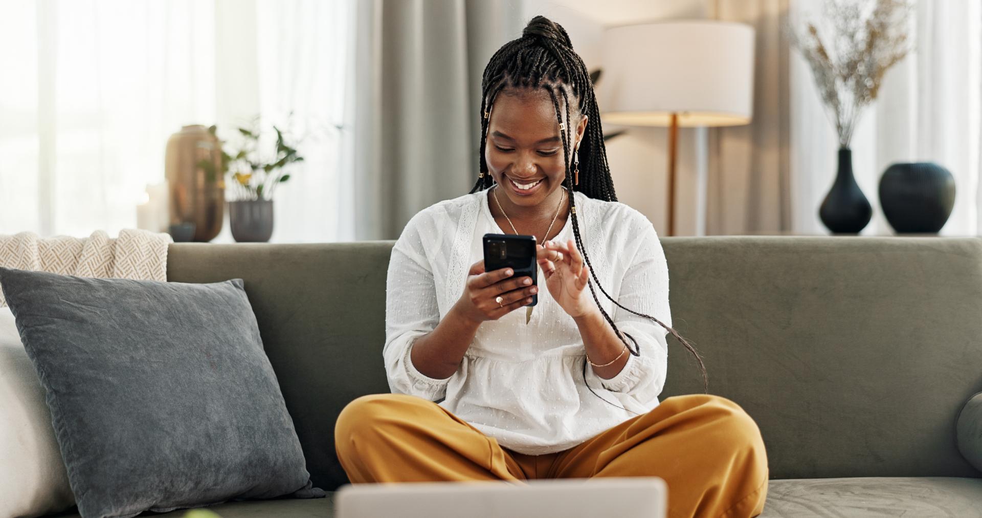 Black woman wearing a white top sitting on a couch smiling at her smartphone