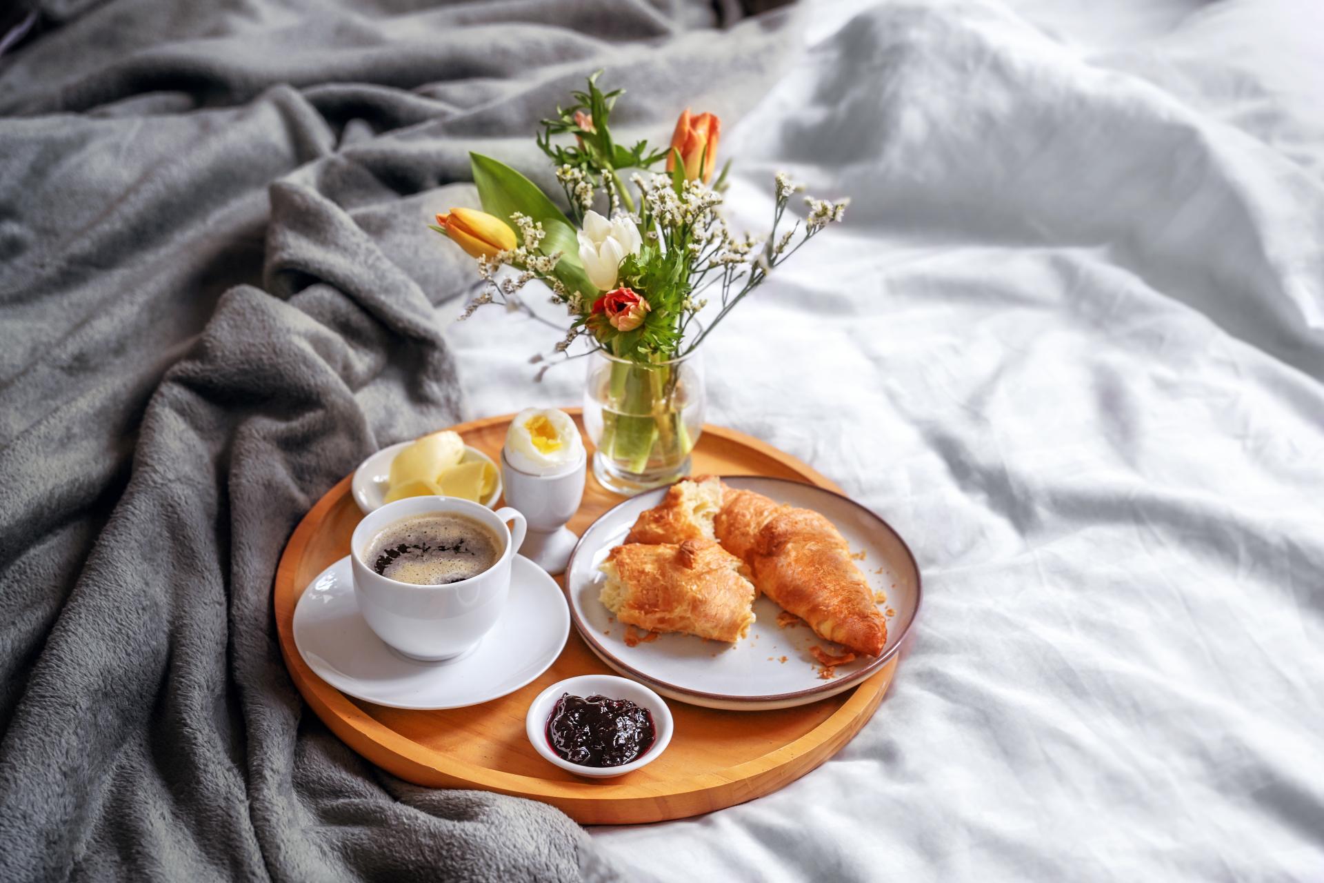 wooden tray on a bed holding coffee croissants jam and an egg