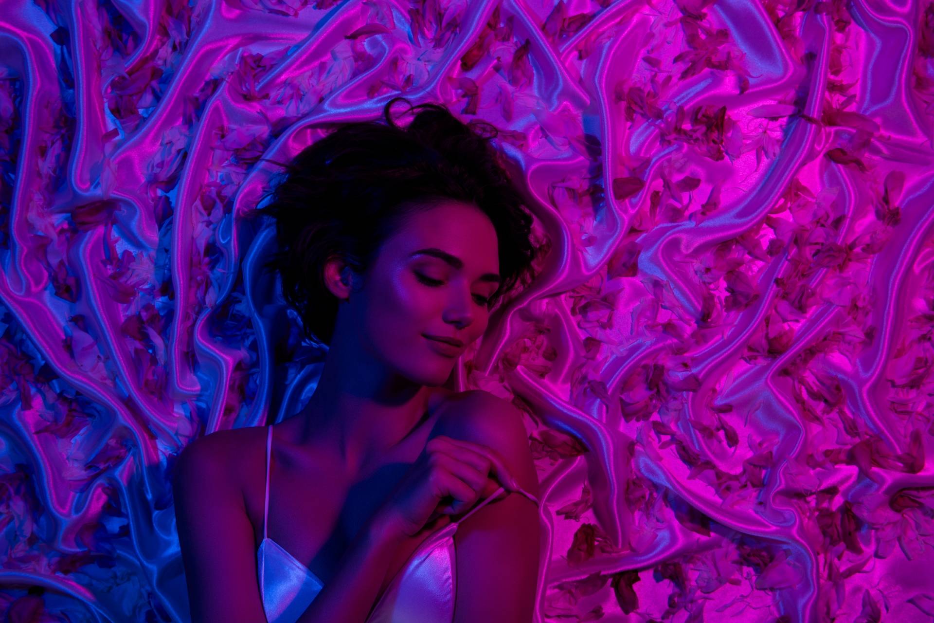 woman in white lingerie on satin sheets covered in petals and neon lighting
