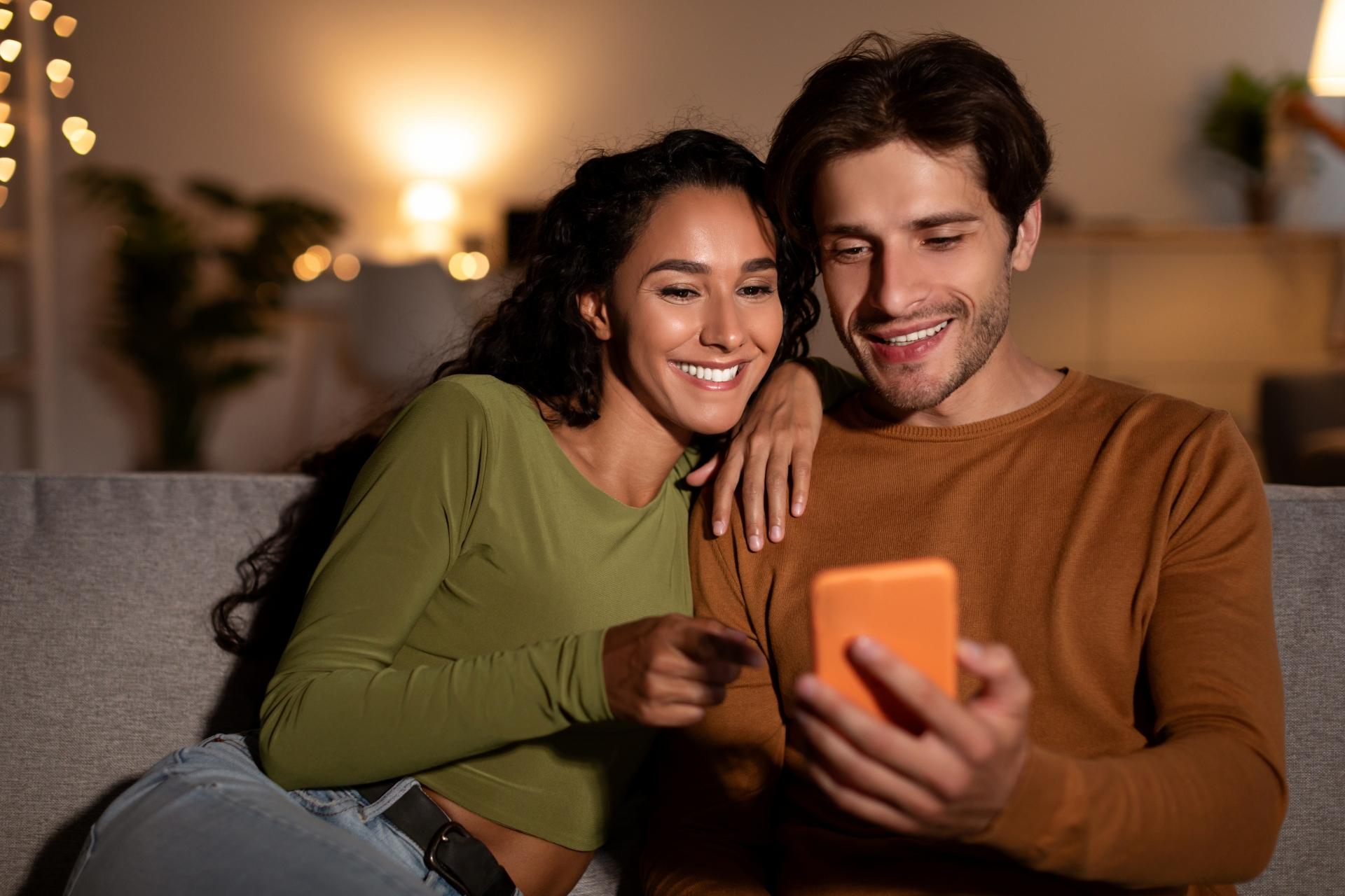 cheerful couple using cellphone sitting on couch at home