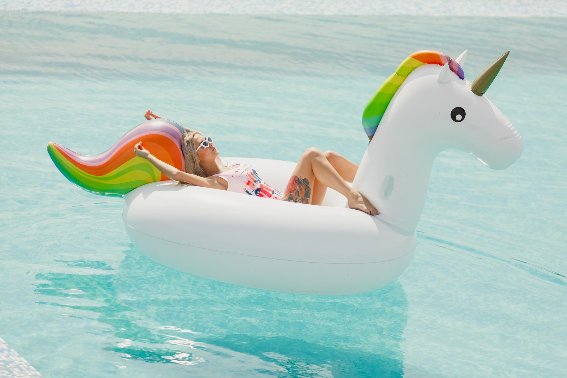 A woman in a pool wearing sunglasses sunbathing on a Unicorn float