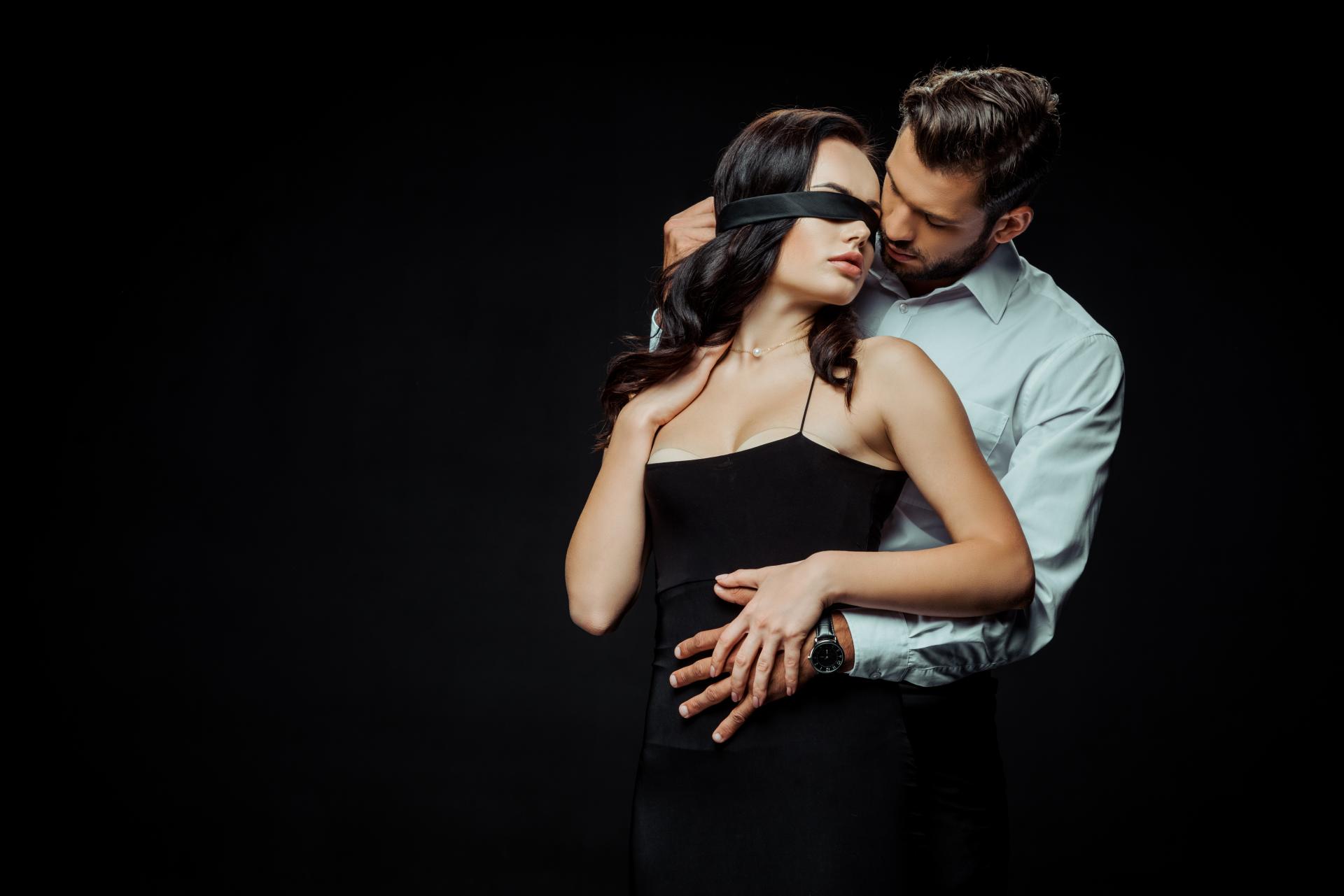 a woman in a black dress and black blindfold with a man holding her from behind