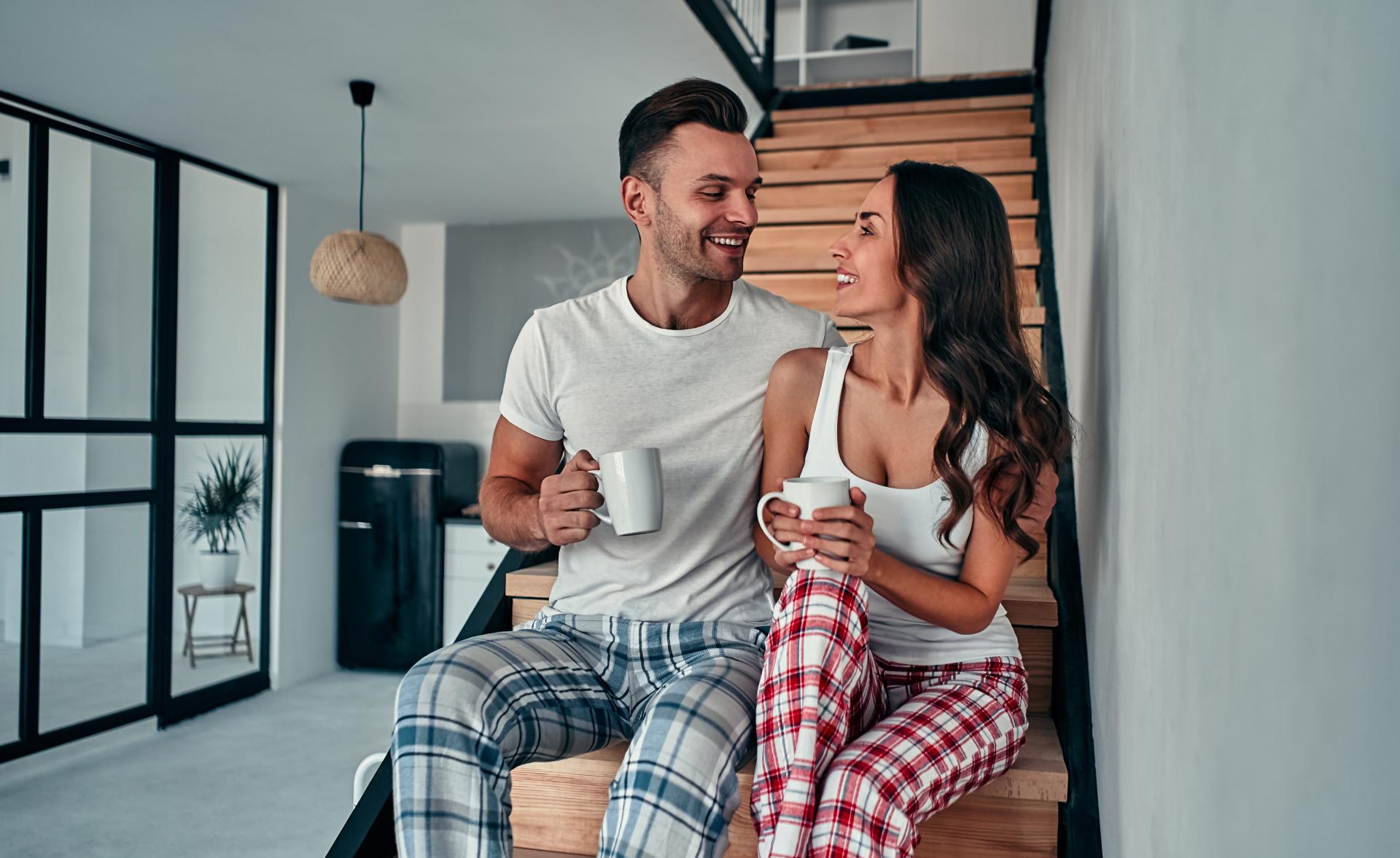 a man and woman sitting on stairs talking in pajamas, each holding a coffee cup