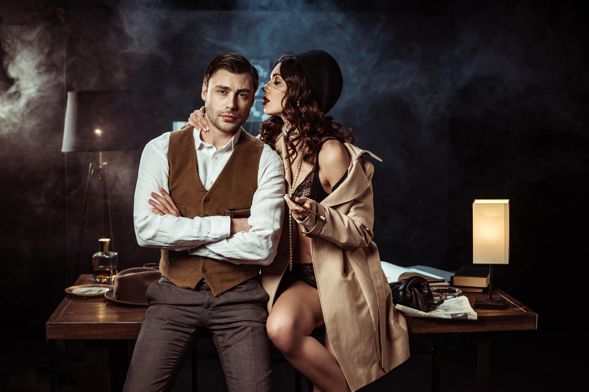 a male detective sitting on a desk with an attractive woman wearing a trench coat and holding a drin