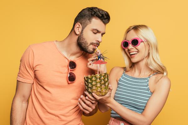 Man in an orange shirt sipping from a pineapple base used as a cup held by a smiling woman over a ye Man in an orange shirt sipping from a pineapple base used as a cup held by a smiling woman over a ye