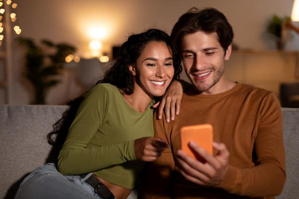 cheerful couple using cellphone sitting on couch at home
