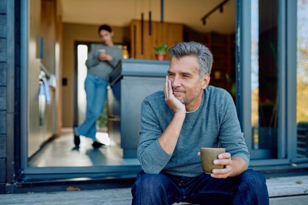man having a hot beverage outside with a woman blurred in the background inside the house