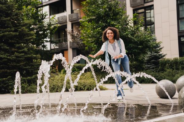 A woman wearing a hat smiling and walking on the edge of a public fountain