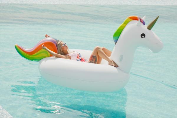 A woman in a pool wearing sunglasses sunbathing on a Unicorn float