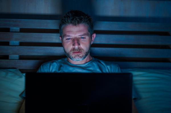 a man wearing a blue shirt appearing exhausted looking at something on his laptop