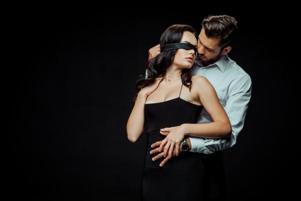 a woman in a black dress and black blindfold with a man holding her from behind