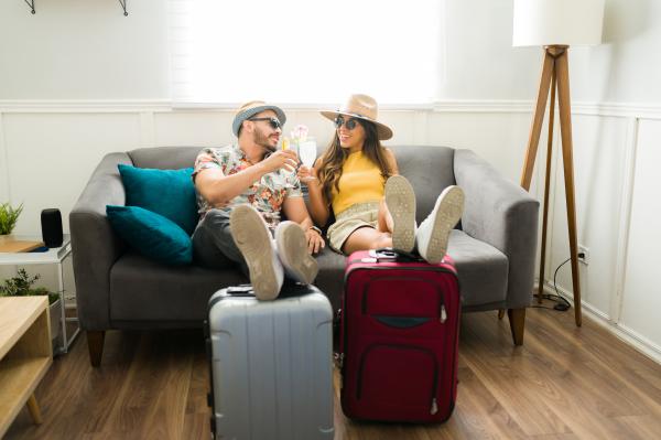 a man and woman wearing hats and sunglasses having a drink on the couch using suitcases as foot rest