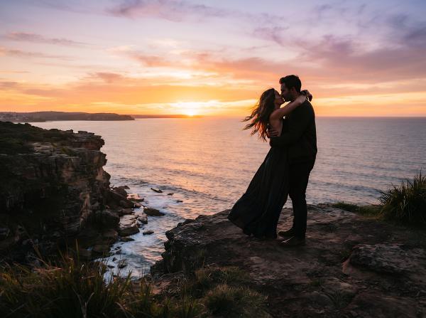 a man and a woman embracing on a cliff with the sun setting in the background